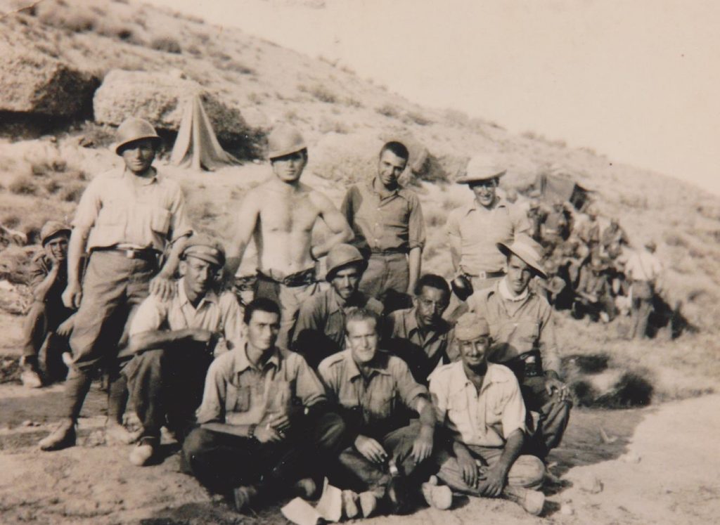 A group of informally dressed soldiers, some wearing helmets, poses for a photo in front of their hillside encampment