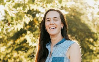Portrait of Lily Rosencrantz, smiling and wearing a sleeveless shirt and jeans, with sun-dappled trees and fields visible in the background